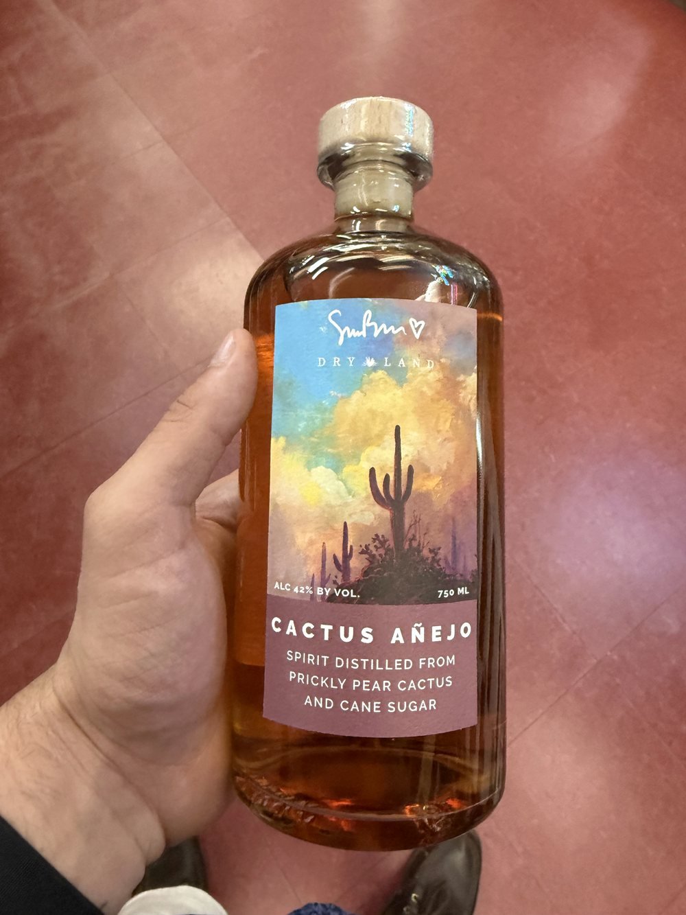 A bottle of Dry Land Distillers Cactus Añejo showing Simon Bull's painted desert label with saguaro cacti at sunset